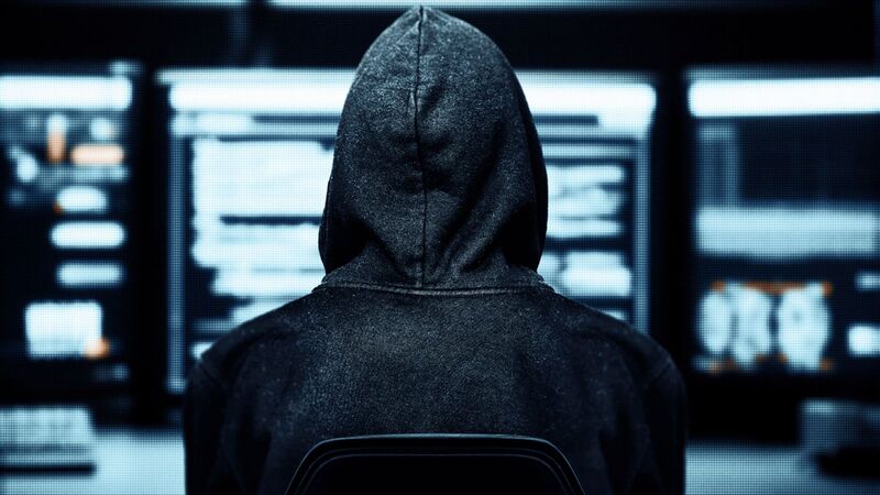 Hacker encapuzado visto de costas em frente a vários monitores, representando ataques cibernéticos, DDoS e ameaças digitais.